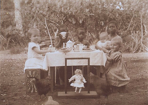 Drei Missionskinder sitzen mit der kamerunischen Nanny und deren zwei Kinder am Tisch im Garten.